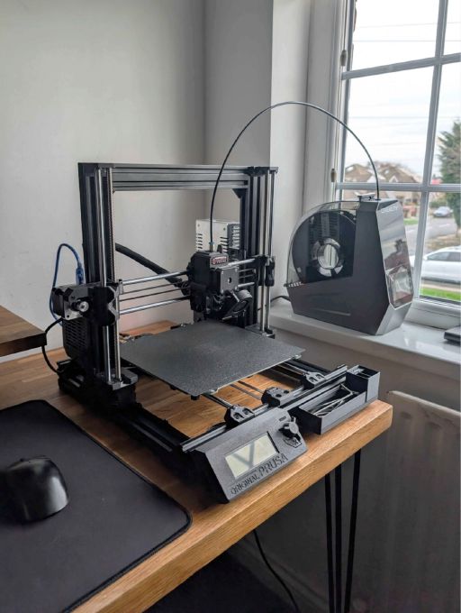 A Prusa MK3 3D printer that has been modified with a black Bear Upgrade frame and a Bondtech extruder. All printed parts are black. The printer stands on a wooden desk next to a window. A spool dryer is on the border of the window. The scene is classy and cosy.