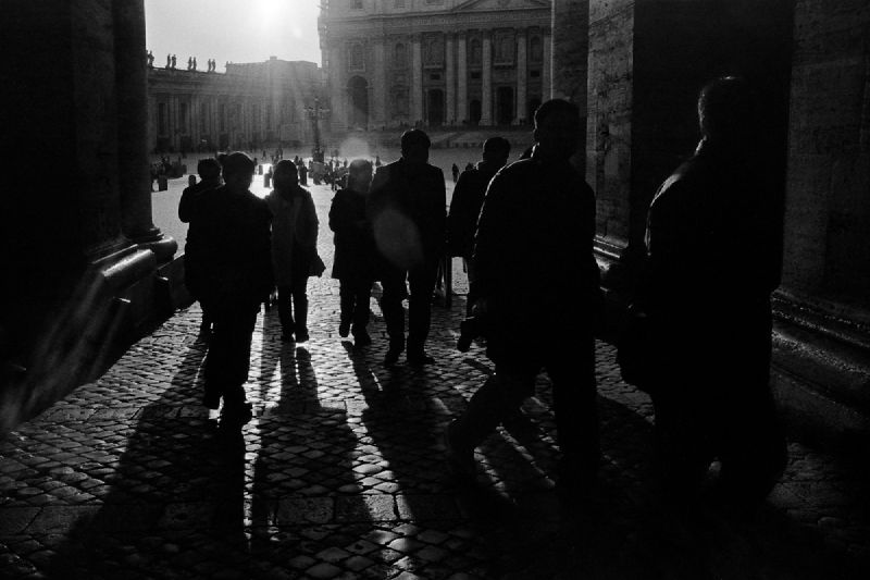 A black-and-white analogue photograph of people walking out of Saint Peter's Square in the Vatican, with the sun creating backlit shadows