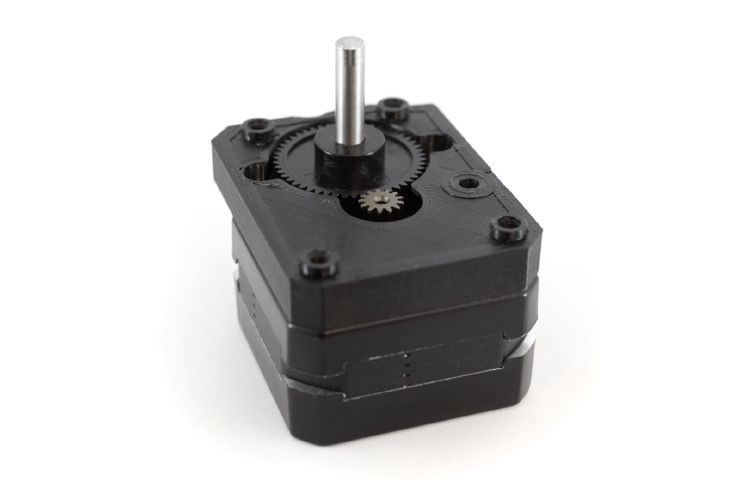 A motor laying on its back with a 3D printed plate attached on top of it. We can see the motor gear in the middle of the plate meshing with another bigger plastic gear