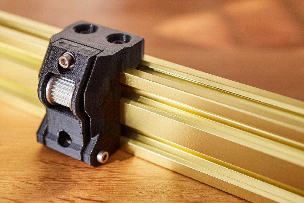 A 3D printed belt tensioner with a large idler bolted onto a gold aluminium extrusion.